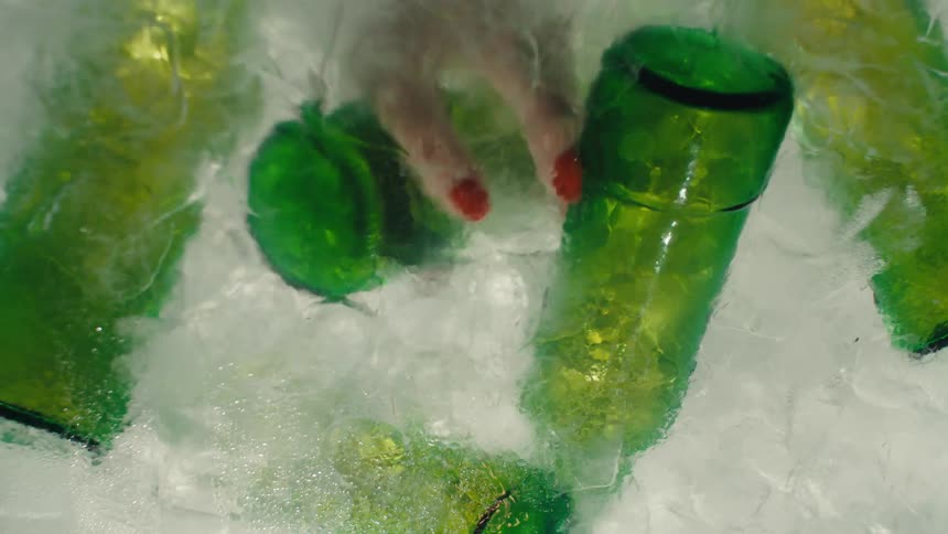 A time-lapse of ice melting around green glass bottles ends with a hand with red nails grabbing a bottle