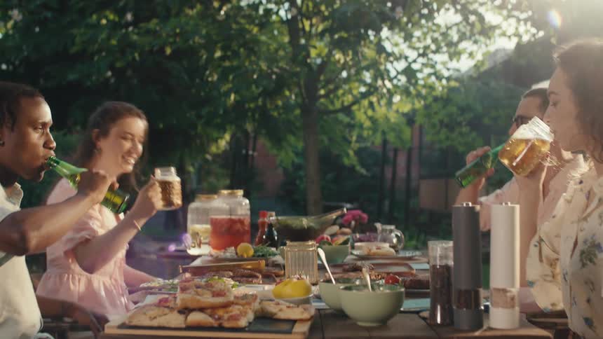 A group of diverse friends joyfully toast with beers and enjoy a festive meal together at a sunny backyard party