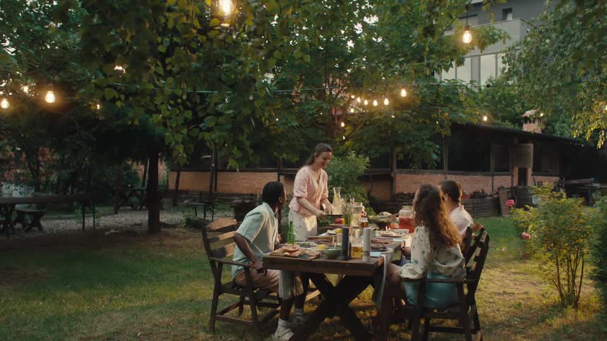 A young woman serves drinks to her diverse group of friends at a cozy backyard dinner party under warm string lights