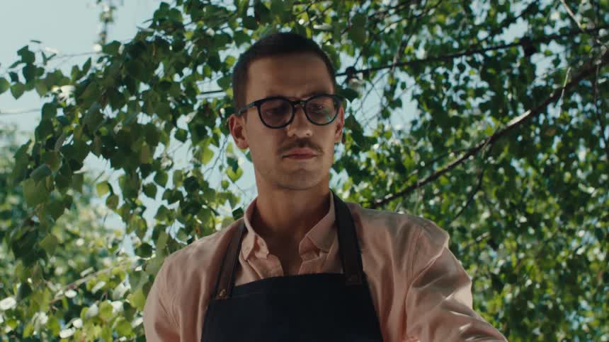 From inside a flaming barbecue, a young man in glasses and an apron lifts the grill grate in slow motion