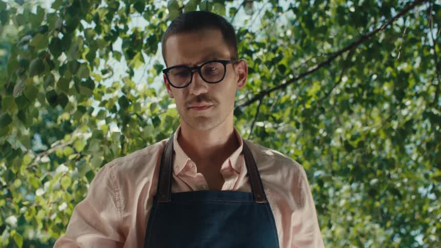From a flaming grill grate, the camera reveals a young man in glasses and an apron ready to barbecue outdoors