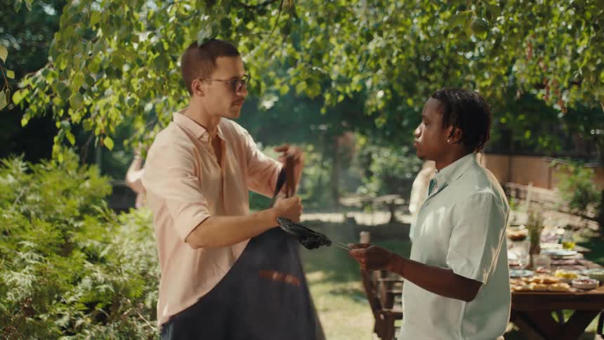 A man hands over his grilling apron to his friend after comically burning the food at a summer BBQ party