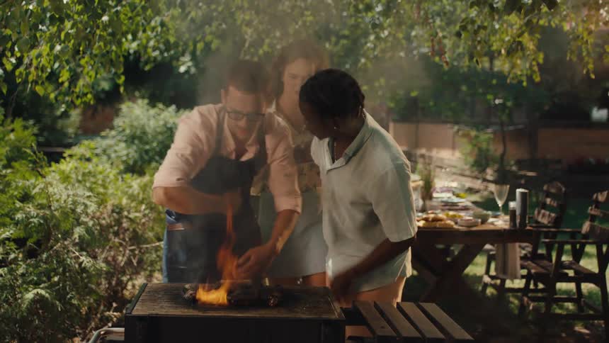 A man in an apron humorously struggles with a smoky, flaming barbecue as his friends look on during a sunny backyard party