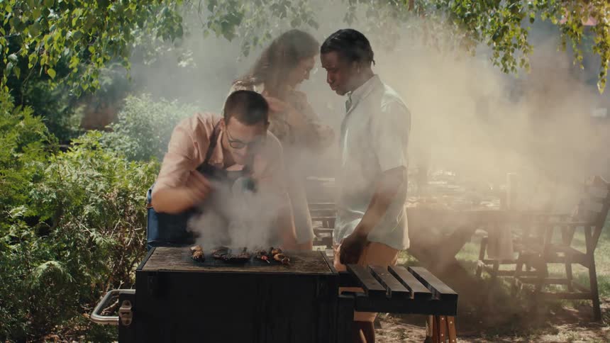 A man in an apron fans the smoke from a barbecue grill while his friends watch during a summer garden party