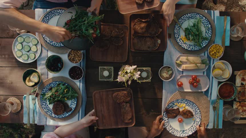 Top-down slow-motion view of friends sharing a delicious meal, passing grilled meats and salads at an outdoor garden party