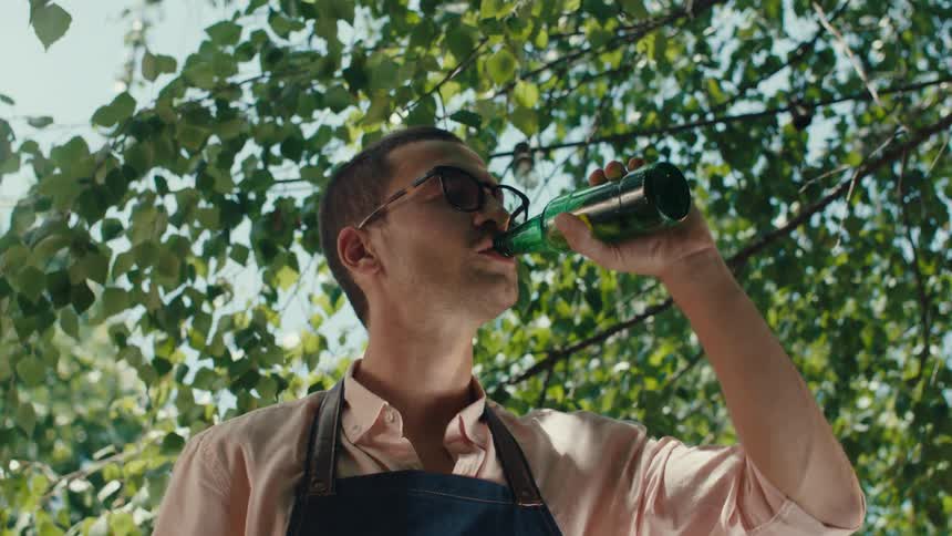 POV from a barbecue grill as a young man wearing glasses and an apron sips a cold beer while cooking outdoors