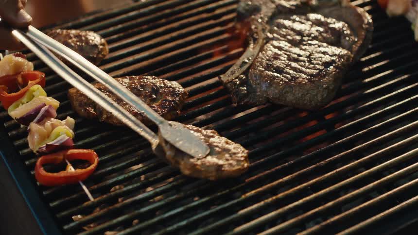 Slow-motion close-up of a T-bone steak, burgers, and vegetable skewers sizzling over the hot flames of a barbecue grill