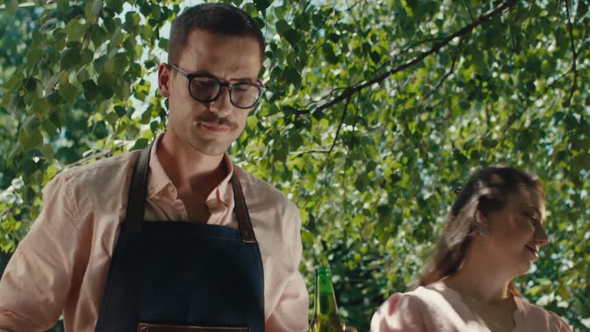 A stylish young man in an apron tends to a barbecue grill as his partner watches lovingly on a sunny day in the garden