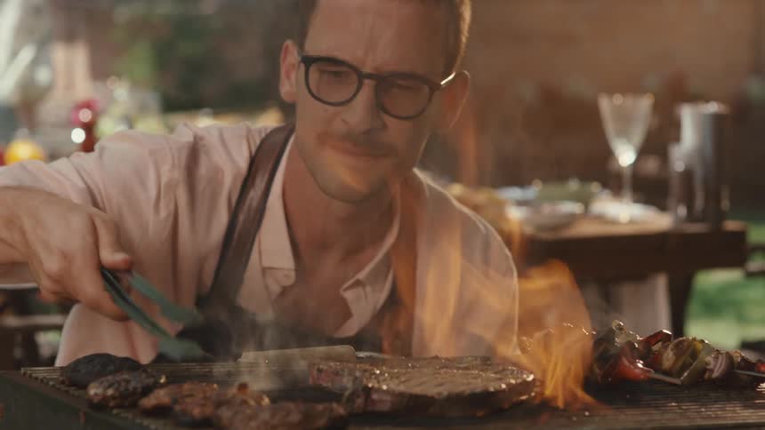 Happy man with glasses and an apron enthusiastically grills steaks and skewers over open flames at a sunny backyard barbecue in slow motion