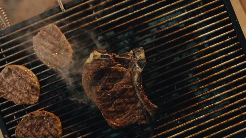 A top-down slow-motion view shows a chef flipping a juicy T-bone steak and grilling burgers on a smoking barbecue