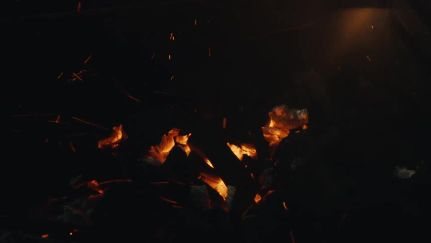 Glowing red hot embers and sparks fly from a fire in slow motion against a dark background