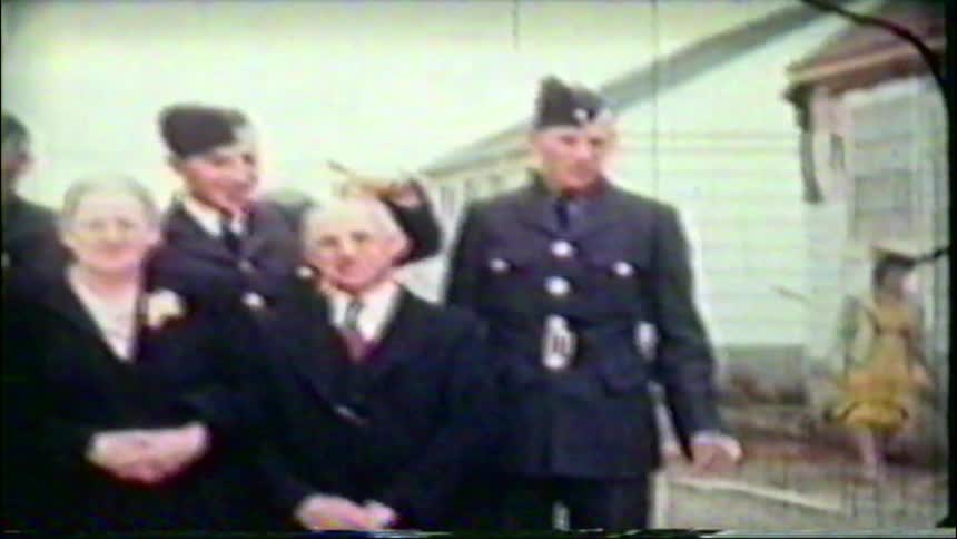 A vintage home movie captures a family posing for a portrait, with two sons in uniform playfully joking around