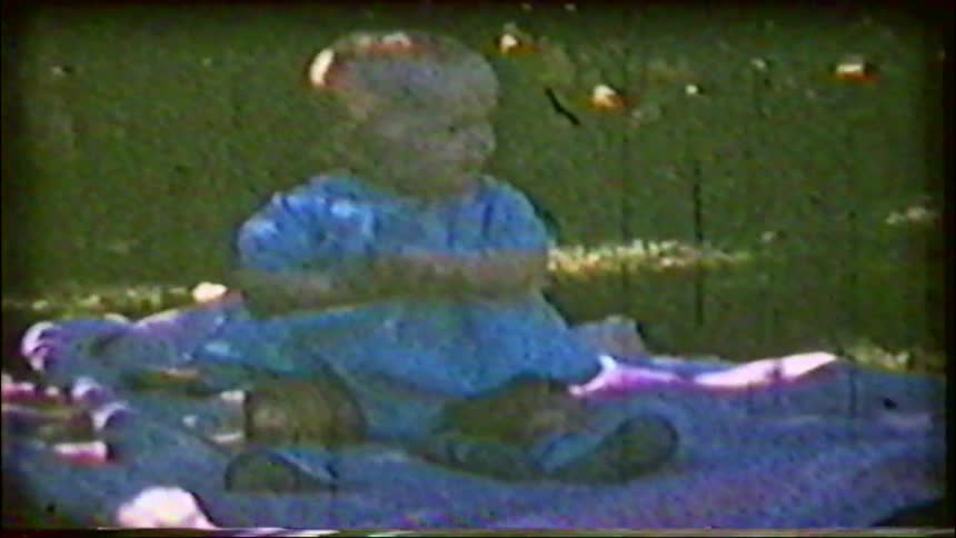 A grainy vintage home movie captures a sweet baby sitting on a colorful blanket on the grass on a sunny day