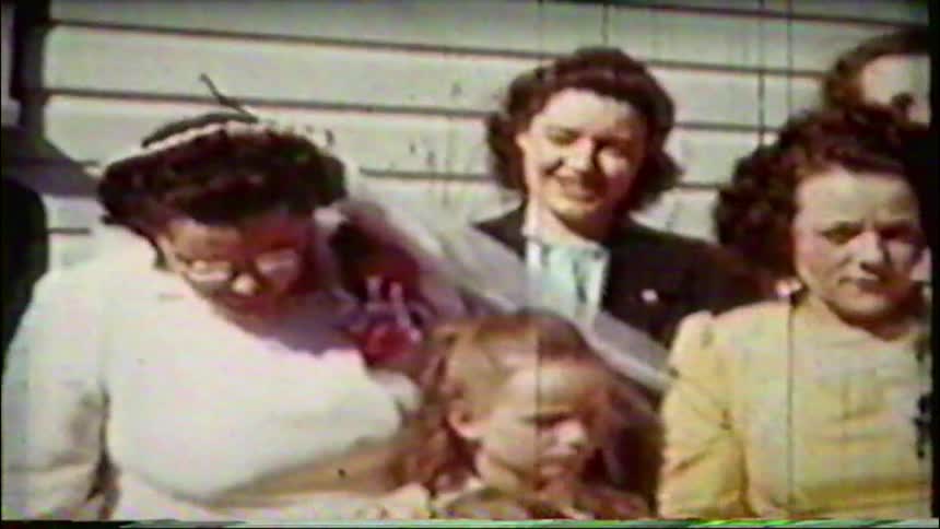 A grainy, vintage home movie from the 1940s captures a joyous family group, including a bride, posing for a picture at an outdoor wedding celebration