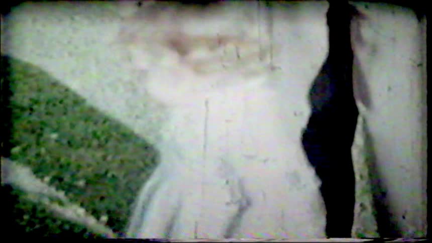 A little blonde girl in a dress stands outside in a grainy, overexposed vintage home movie from the mid-20th century