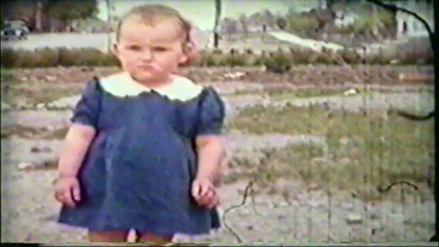 A vintage home movie captures an adorable toddler girl in a blue dress, whose serious face breaks into a happy smile as she begins to walk