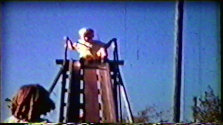 A young child hesitantly slides down a tall playground slide into their mother's waiting arms in this nostalgic vintage film