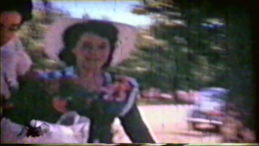 A vintage home movie captures a smiling woman in a wide-brimmed hat standing outdoors on a sunny day
