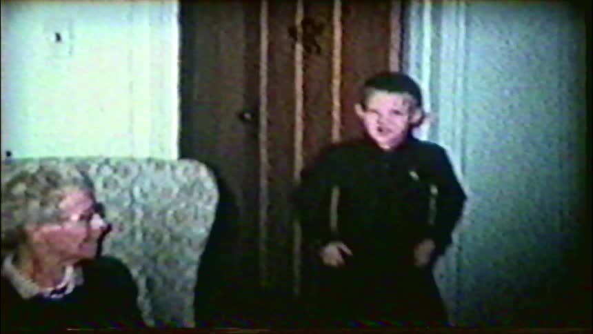 A vintage home movie captures a young boy posing for the camera next to his grandmother in their living room