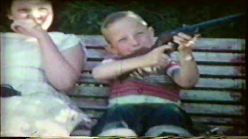 A vintage home movie from the 1950s shows three siblings laughing and making silly faces while sitting on a porch swing, as the youngest boy aims a toy gun