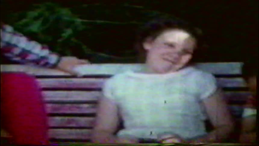 Vintage home movie captures three children sitting together on an outdoor bench, with one young boy smiling as he aims a toy rifle