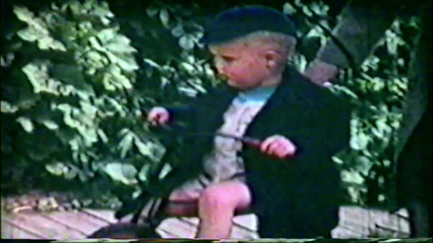 A vintage home movie shows a cute toddler boy in a cap and jacket sitting on his tricycle in the backyard