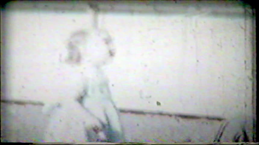An adorable little girl with blonde curly hair stands in a backyard in this grainy, vintage 8mm home movie footage