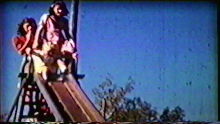 Three young sisters sit at the top of a playground slide on a sunny day in this nostalgic vintage home movie