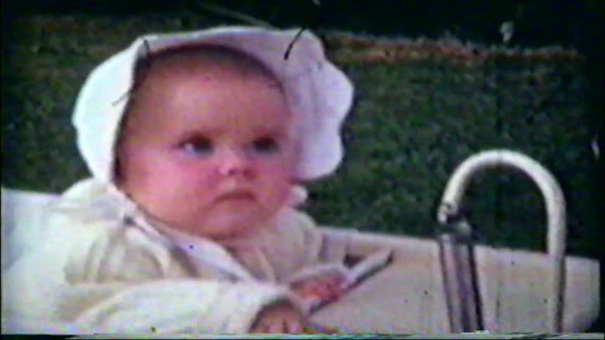 A vintage home movie captures a baby in a white bonnet sitting in a stroller, initially looking serious before breaking into a joyful smile