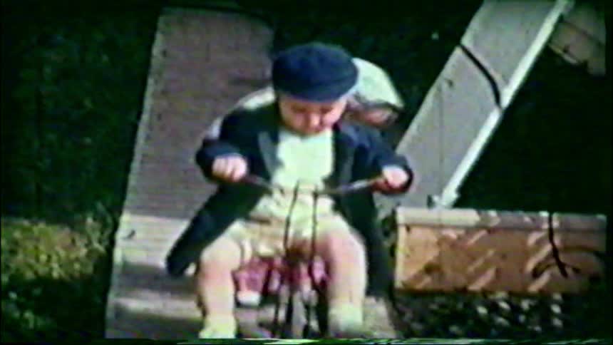 A nostalgic home movie from the 1950s shows a little boy on a tricycle and his sister playing in the backyard