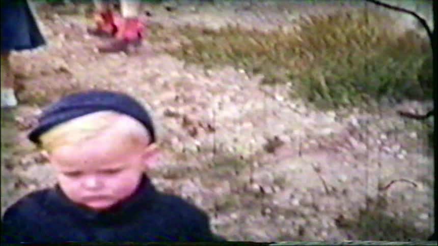 A toddler with bright blonde hair and a dark cap walks outdoors in a grainy vintage home movie