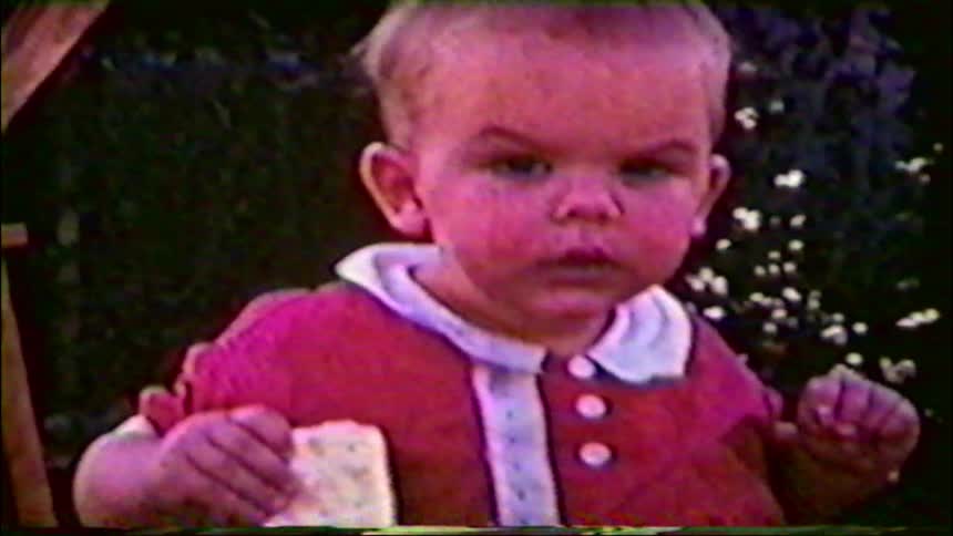 A close-up of a cute baby in a red outfit eating a snack in a garden from a vintage home movie