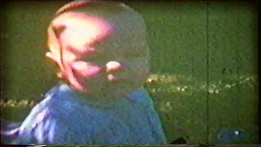 A vintage home movie captures a close-up of a cute baby sitting in the grass on a sunny day