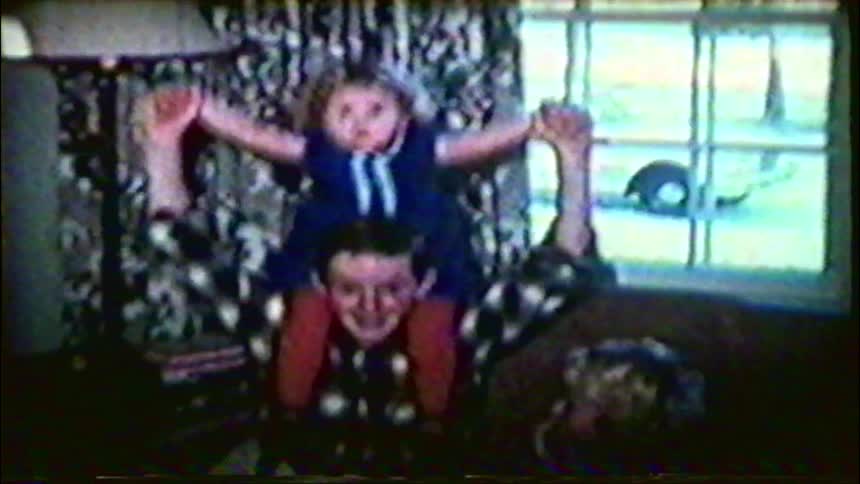 A father and his two young daughters pose playfully for a vintage home movie in their living room