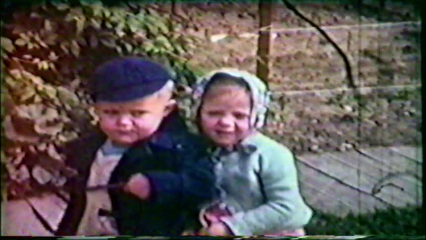 Vintage 8mm home movie footage shows a little boy and a teary-eyed girl sitting close together in a garden