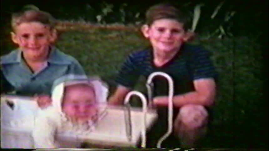 Two young brothers smile for the camera next to their baby sibling in a vintage rocker in the backyard