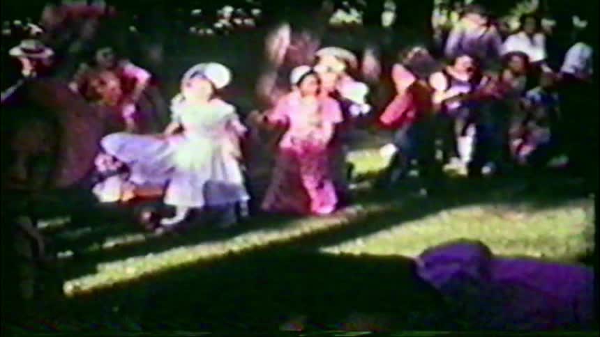 Children dressed in colorful, old-fashioned costumes gather for an outdoor event in a grainy vintage home movie