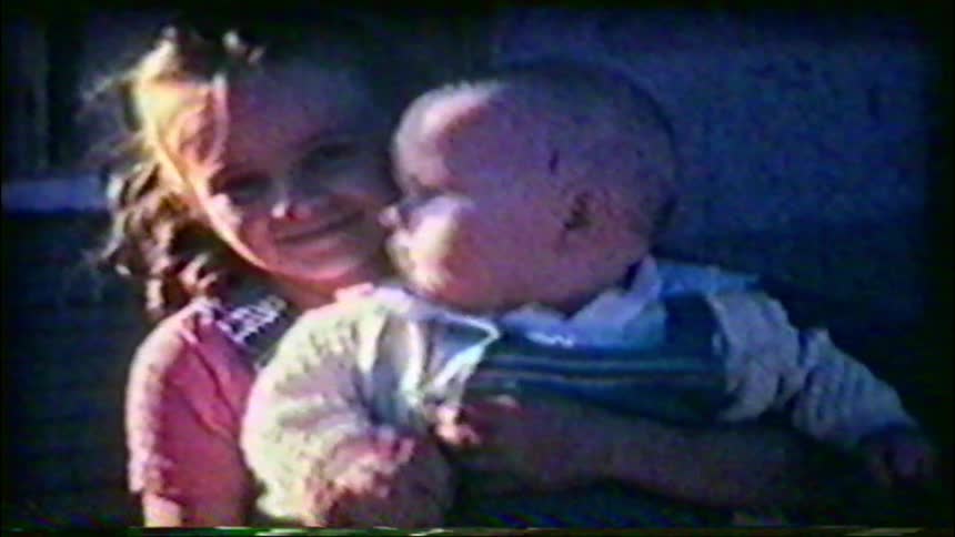 Vintage home movie footage captures a sweet moment of a little girl smiling as she holds her baby brother