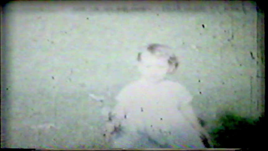 A grainy and faded vintage home movie captures a small toddler in a dress standing outdoors
