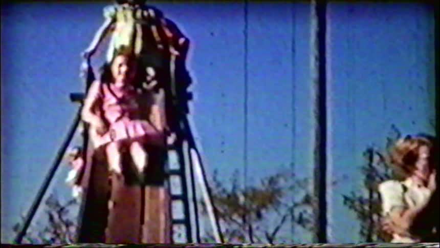 Vintage footage shows a joyful girl in a pink dress going down a tall slide at the playground on a sunny day