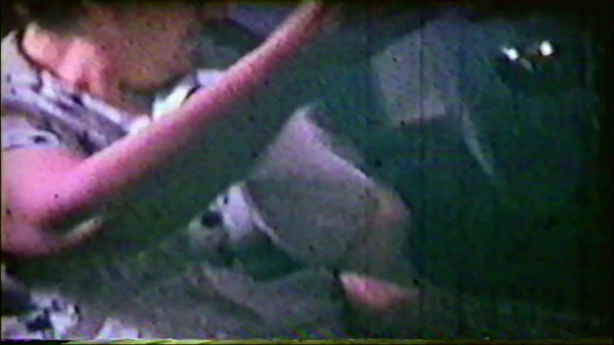 A grainy vintage home movie captures an elderly woman unwrapping a gift, which appears to be a photo album or scrapbook