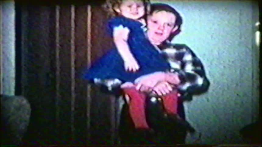 A nostalgic home movie captures a boy carrying his little sister into the living room to pose for the camera