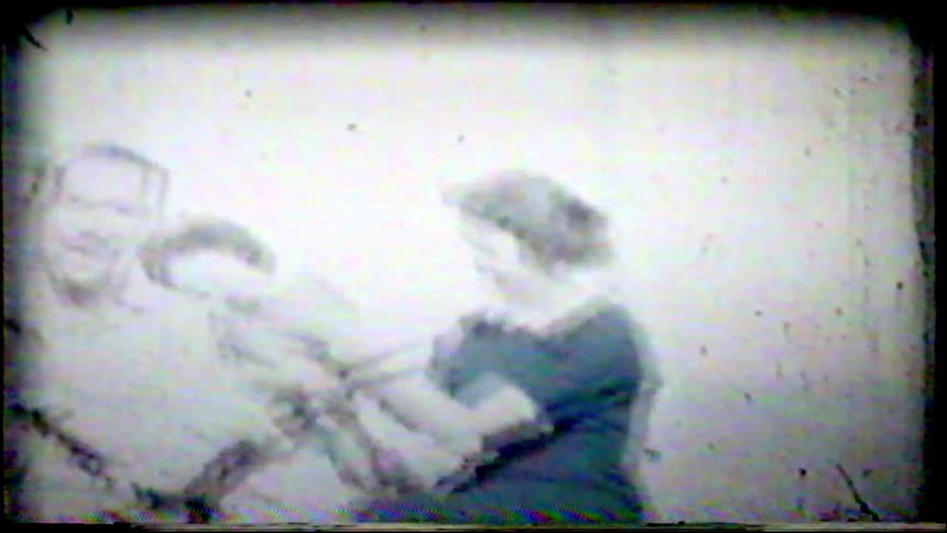 A father plays the violin with his children and wife sitting beside him in a grainy black and white vintage home movie