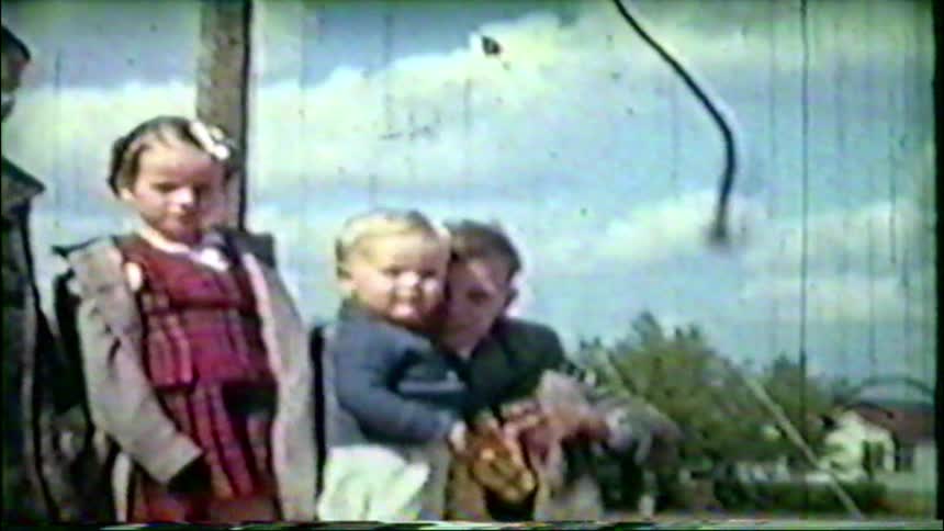A vintage home movie from the 1950s shows a father and his young children posing together outside