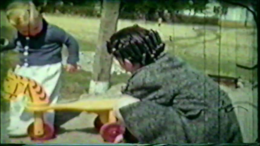 Grainy vintage home movie footage shows two young children playing with toys in a suburban backyard