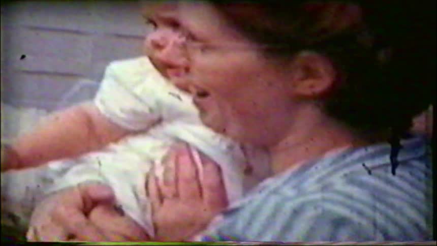 Vintage 8mm home movie footage shows a loving mother in glasses holding and cuddling her baby outdoors