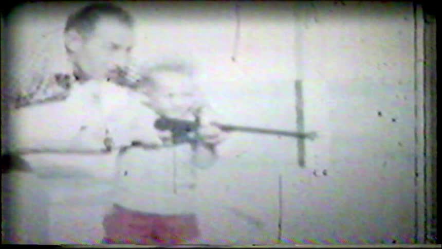 Vintage 8mm film captures a father teaching his young son how to hold and aim a toy Tommy gun in the backyard