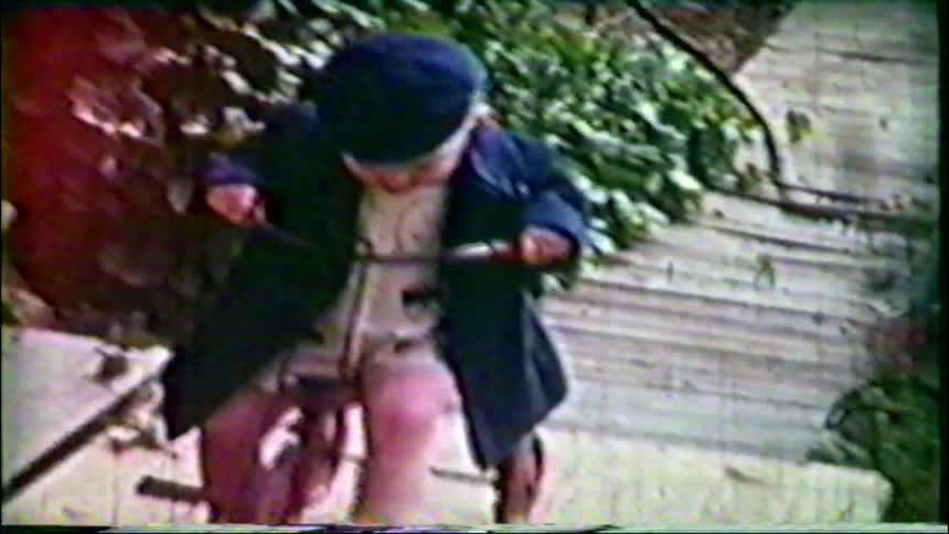 Archival footage captures a toddler wearing a coat and cap sitting on a tricycle in a garden
