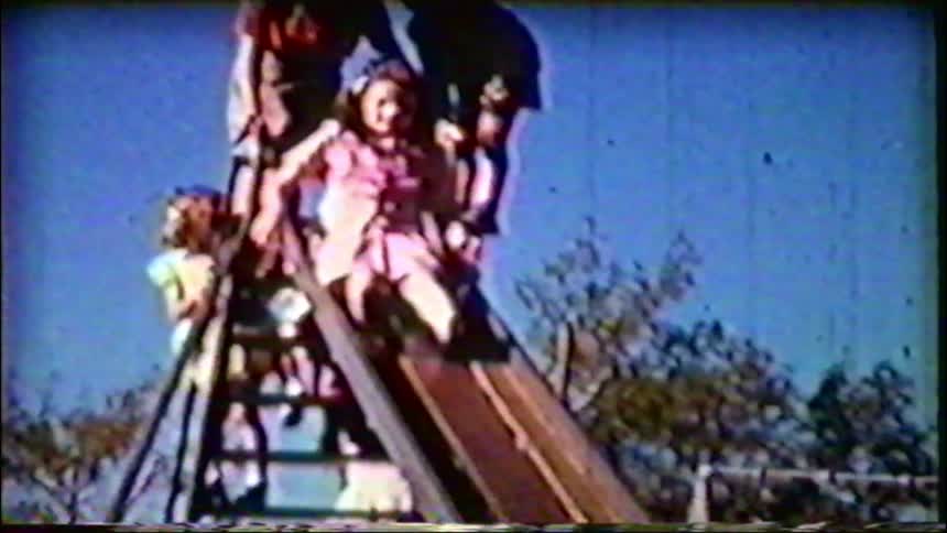 Grainy, nostalgic home movie captures a young girl sliding down a playground slide on a sunny day