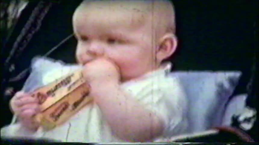 A vintage home movie from the 1960s shows a close-up of a baby, followed by a shot of the baby sitting in a pram outdoors